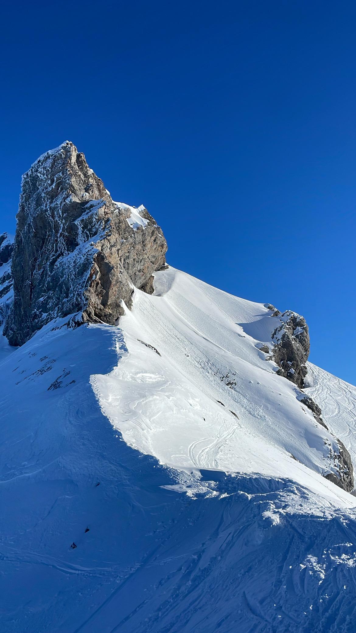 A rocky summit and its ridge under fresh snow, against a cloudless blue sky.